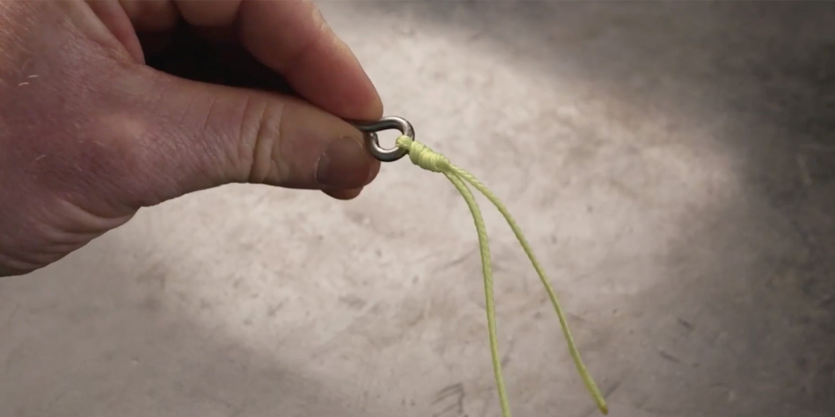 A hand holding a fishing hook attached to a yellow fishing line, which is looped and knotted.